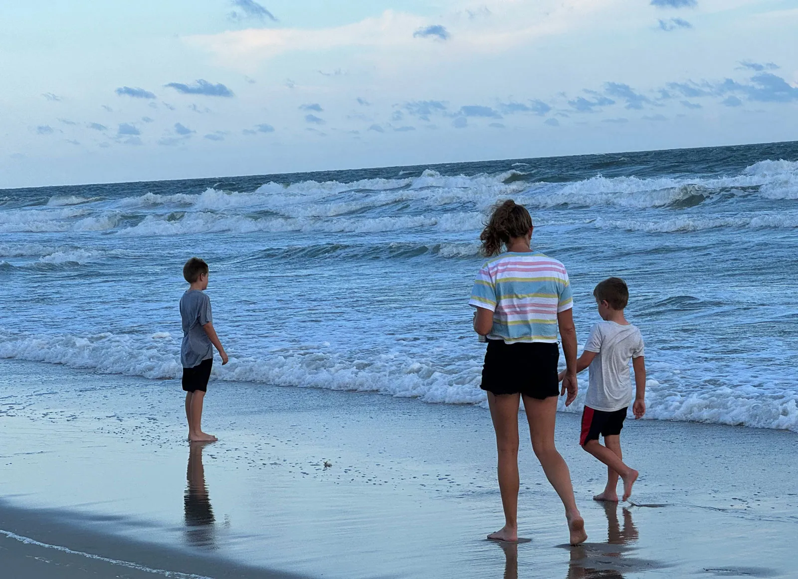 Three children wading at the surf line at dusk, seen from behind